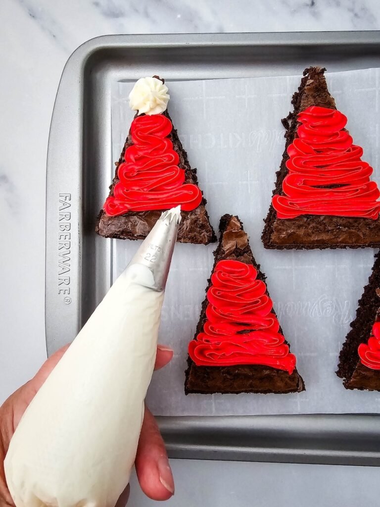 Piping white frosting onto red iced brownies to create Santa hat trim and pom-pom
