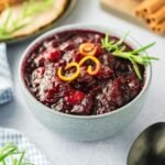 a bowl of homemade cranberry sauce garnished with orange zest and rosemary, ready to serve for Thanksgiving dinner.