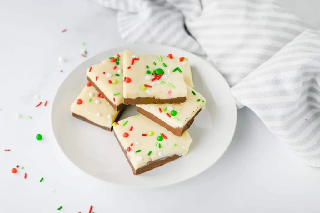 Plate of layered Christmas chocolate fudge squares with white chocolate tops and red, green, and white sprinkles, ready to serve.
