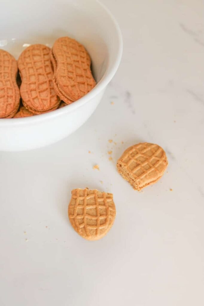 Close-up of white bowl with two cookies cut in half on a marble surface, showing the first step of making Nutter Butter acorns.