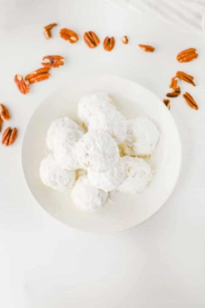 Overhead view of pecan snowball cookies dusted in powdered sugar on a white plate with scattered pecans on the table.