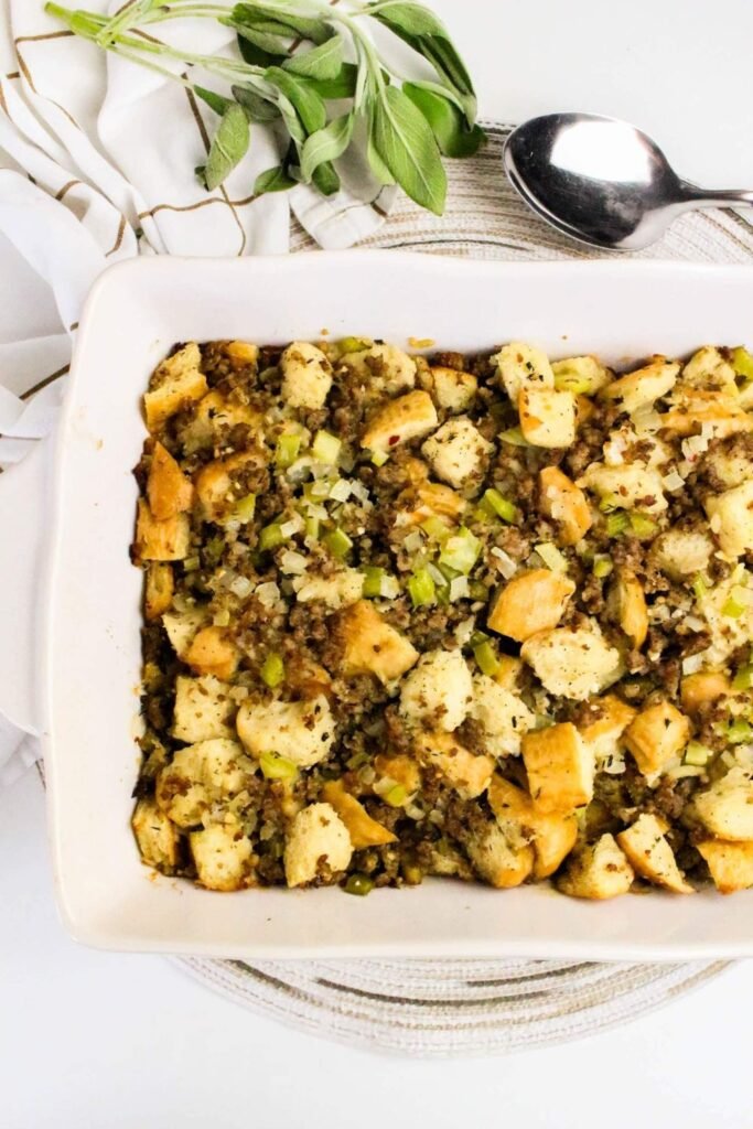 Baked sage and sausage herb stuffing in a white casserole dish with golden bread cubes, celery, and fresh sage garnish.