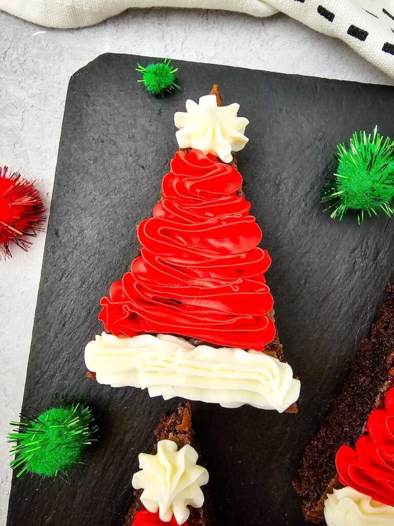 “cose-up of a Santa Hat Brownie decorated with red and white frosting on a slate board with holiday pom-poms.