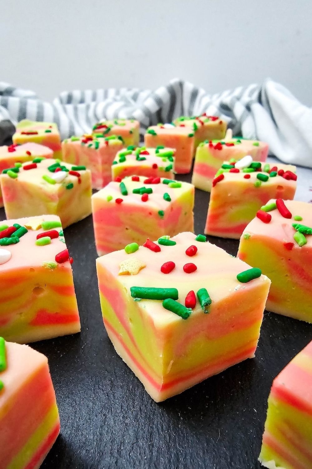 Close-up of creamy swirled Christmas fudge squares topped with red and green sprinkles, arranged on a black slate for a festive holiday dessert.
