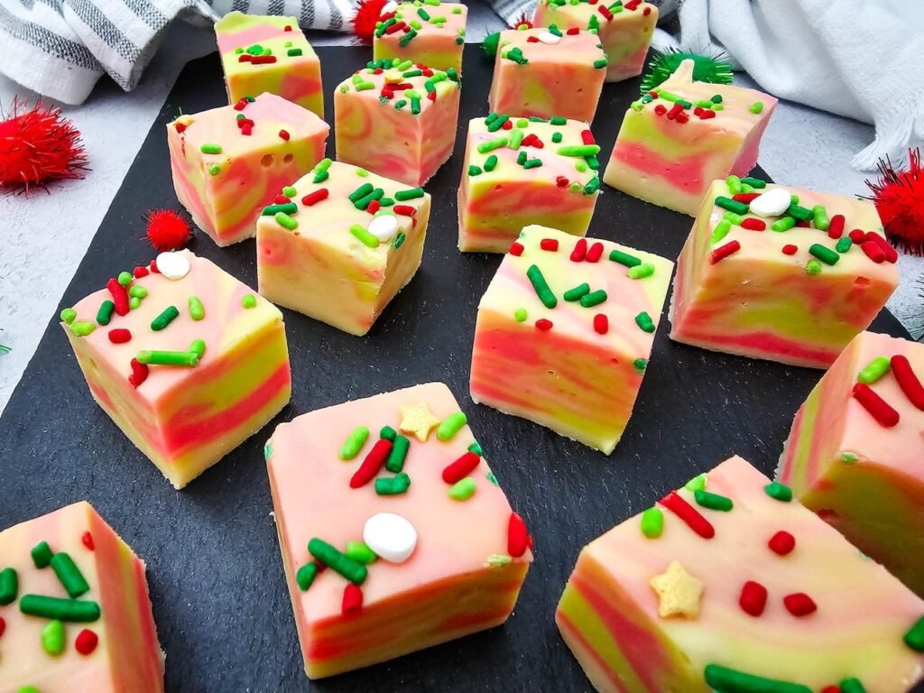 Squares of creamy swirled Christmas fudge with red and green sprinkles arranged on a black slate serving tray.