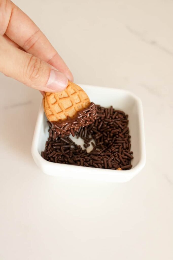 Hand dipping a chocolate-covered half into a small dish of chocolate sprinkles, showing step four in the process.