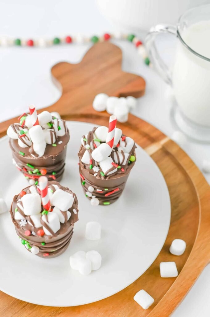 Three homemade hot chocolate sticks decorated with mini marshmallows and red, green, and white Christmas sprinkles on a white plate beside a glass of milk.