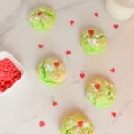 Bright green Christmas cookies topped with red heart sprinkles on a marble counter, surrounded by extra hearts and a jar of milk.
