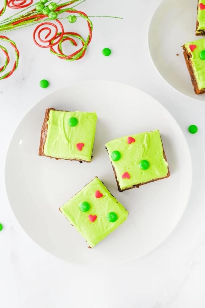 Overhead view of three Grinch Brownies with green frosting, red heart sprinkles, and green M&Ms on a white plate, surrounded by festive red and green holiday ribbon.
