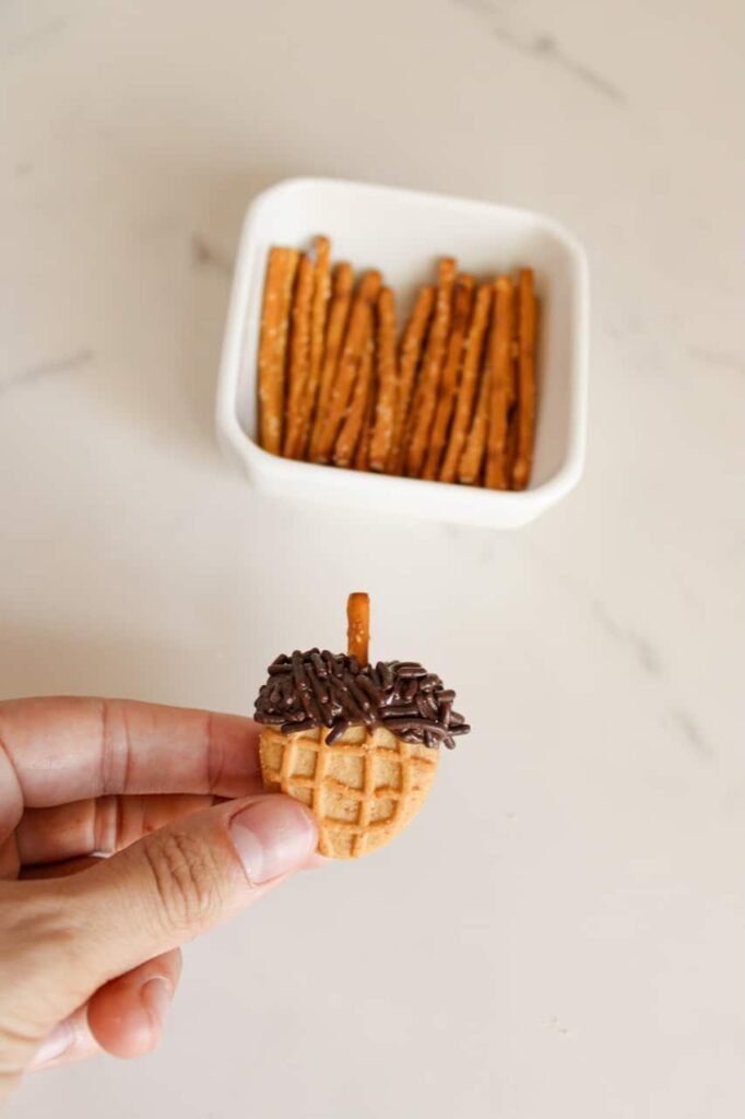 Hand holding a finished cookie with chocolate sprinkles on top and a pretzel stick stem, showing step five of the recipe process with a bowl of pretzels in the background.