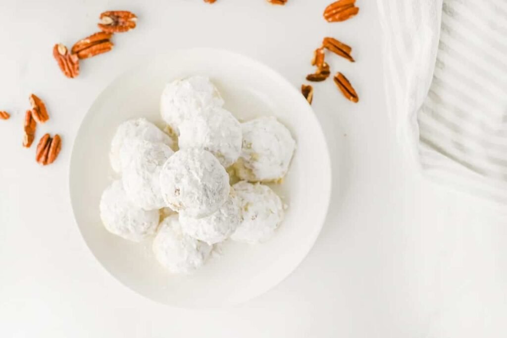 Overhead flat lay of pecan snowball cookies covered in powdered sugar on a white plate, surrounded by scattered pecans and a striped kitchen towel.