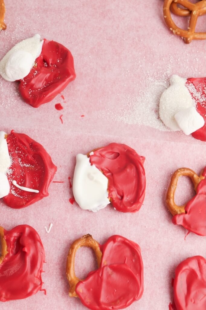 Pretzels dipped in red and white candy melts with mini marshmallows and sugar sprinkles added to form Santa hat treats.