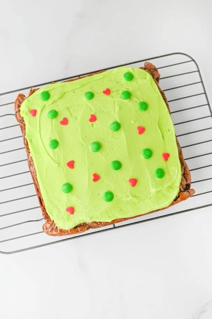 Finished desserttopped with bright green frosting, red heart sprinkles, and green M&Ms cooling on a wire rack, ready to slice and serve for Christmas.