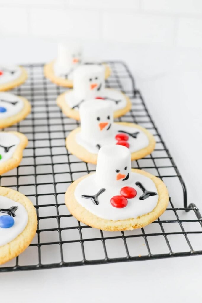 Fully decorated melted snowman cookies with marshmallow heads, icing faces, and colorful candy buttons cooling on a wire rack.