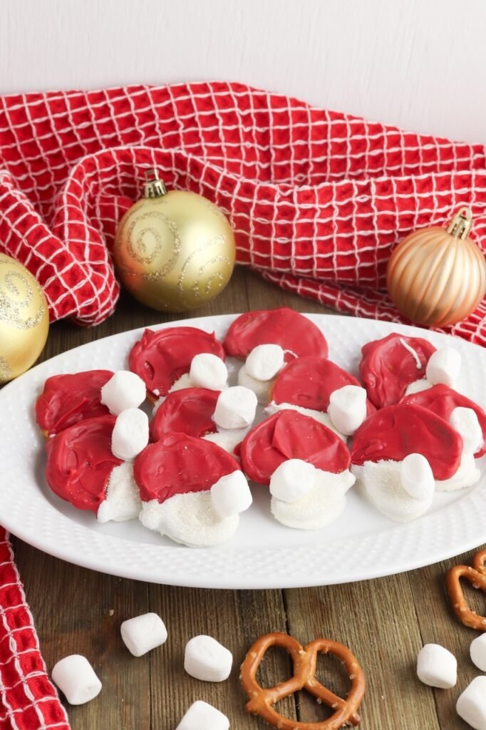 A white plate filled with red and white Pretzel Santa Hats topped with mini marshmallows, surrounded by gold ornaments and a red holiday towel.
