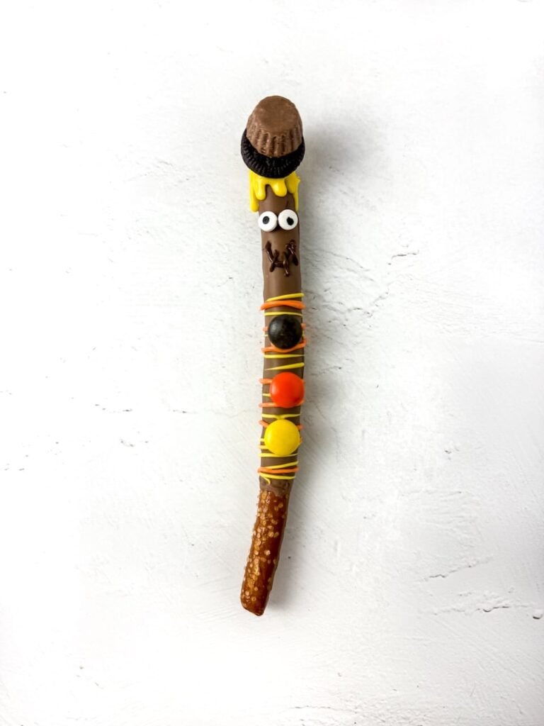 Fully decorated scarecrow pretzel rod coated in chocolate and topped with a mini peanut butter cup and Oreo hat. The pretzel features yellow candy melt “straw” hair, candy eyes, a chocolate smile, and Reese’s Pieces buttons in orange, yellow, and brown. The no-bake fall treat is displayed on a white background.