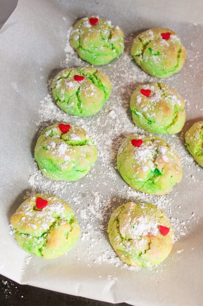 Freshly baked green holiday cookies topped with red heart sprinkles and dusted with powdered sugar on parchment paper.
