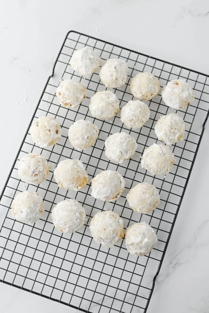 Freshly baked pecan snowball cookies coated in powdered sugar cooling on a black wire rack after baking.