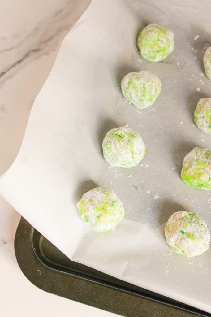 Green cookie dough balls coated in powdered sugar arranged on a parchment-lined baking sheet, ready to bake.