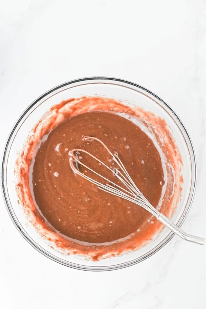 Mixing chocolate brownie batter in a glass bowl with a metal whisk during the first step of making Grinch Brownies.