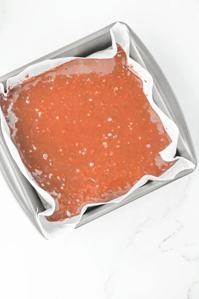 Unbaked batter poured into a square metal pan lined with parchment paper, ready to go into the oven.