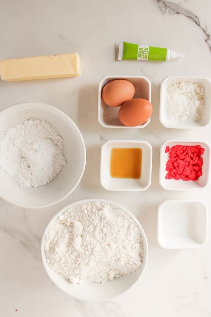 Overhead view of ingredients for green holiday cookies made with cake mix, butter, eggs, powdered sugar, and red heart sprinkles.