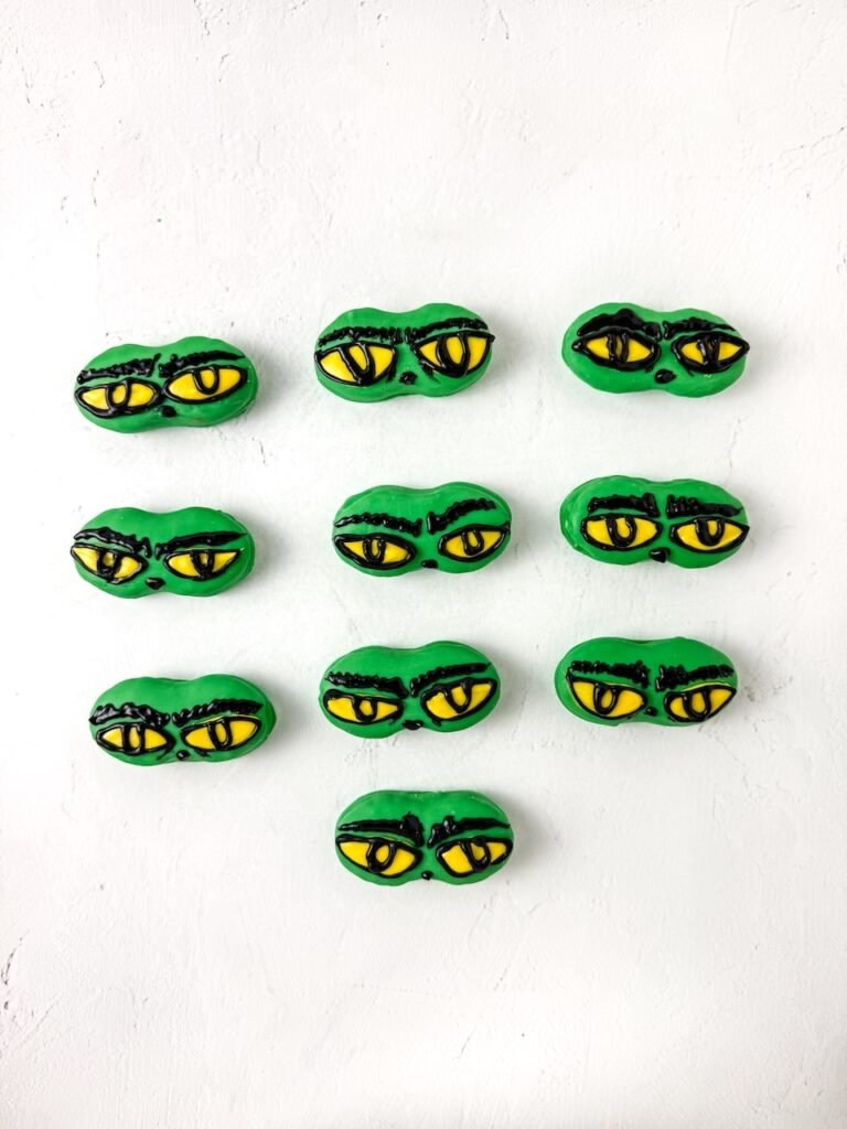 Batch of finished Grinch Nutter Butter cookies decorated with yellow eyes and black eyebrows arranged on a white background.