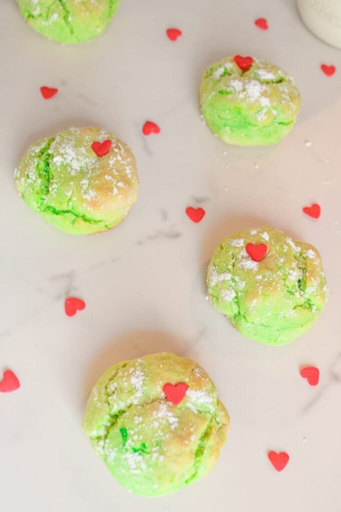 Overhead view of green Grinch Cookies topped with red heart sprinkles and dusted with powdered sugar on a marble surface.