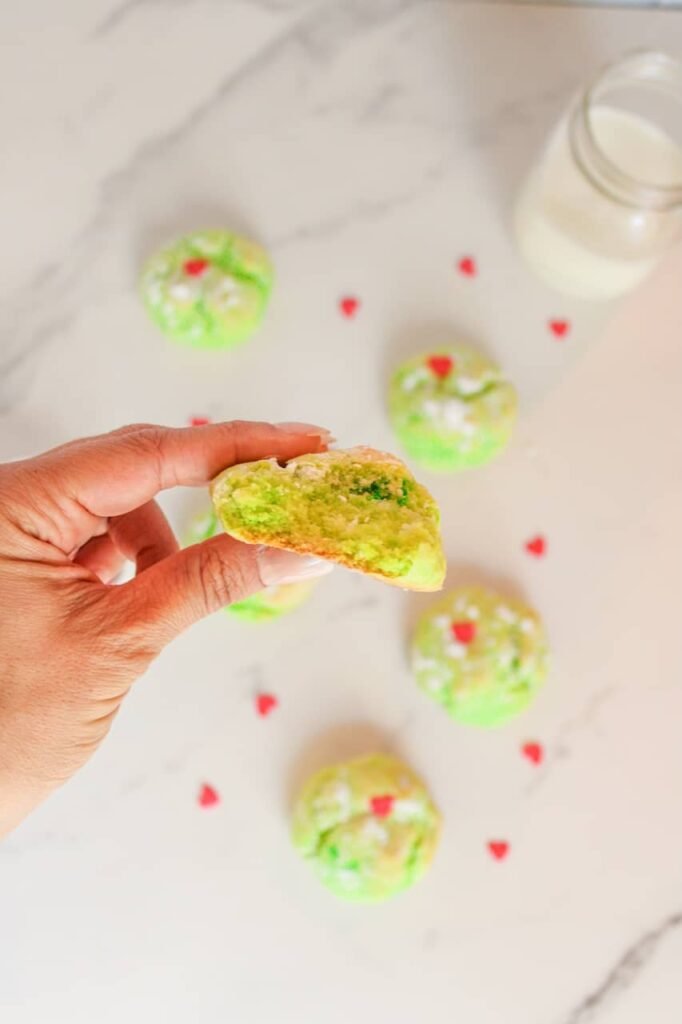 Close-up of a hand holding a soft Grinch Cookie with a bite taken out, showing its chewy green center and festive red heart decoration.