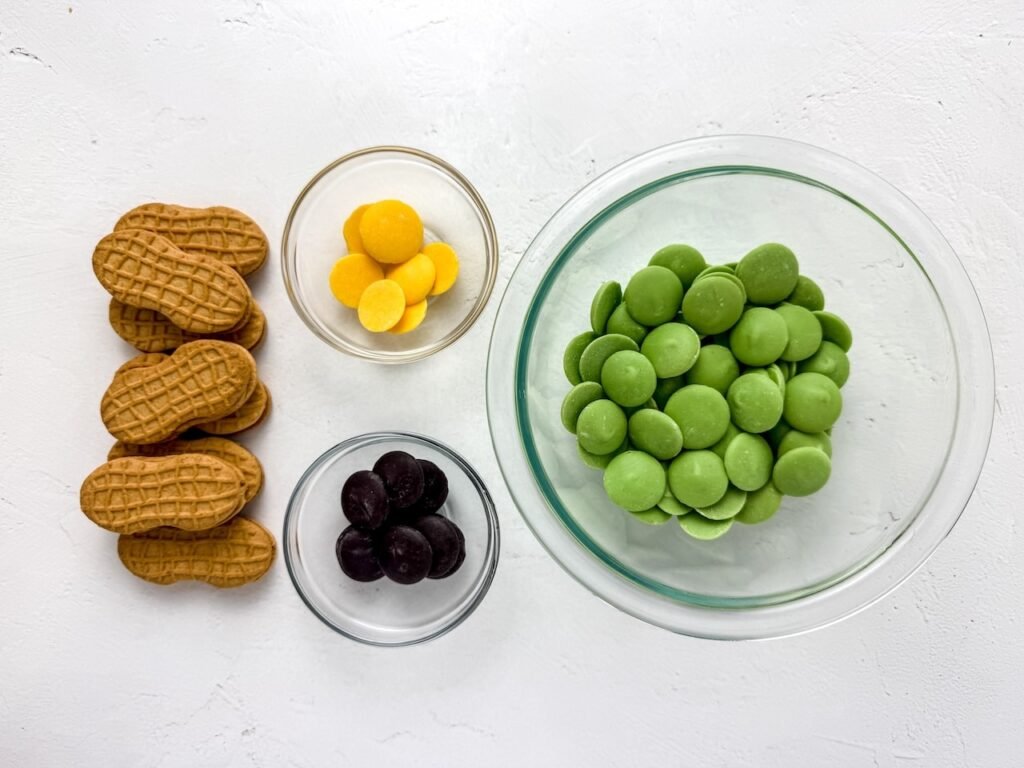 ingredients including biscuits, green candy melts, yellow candy melts, and black candy melts arranged in bowls.
