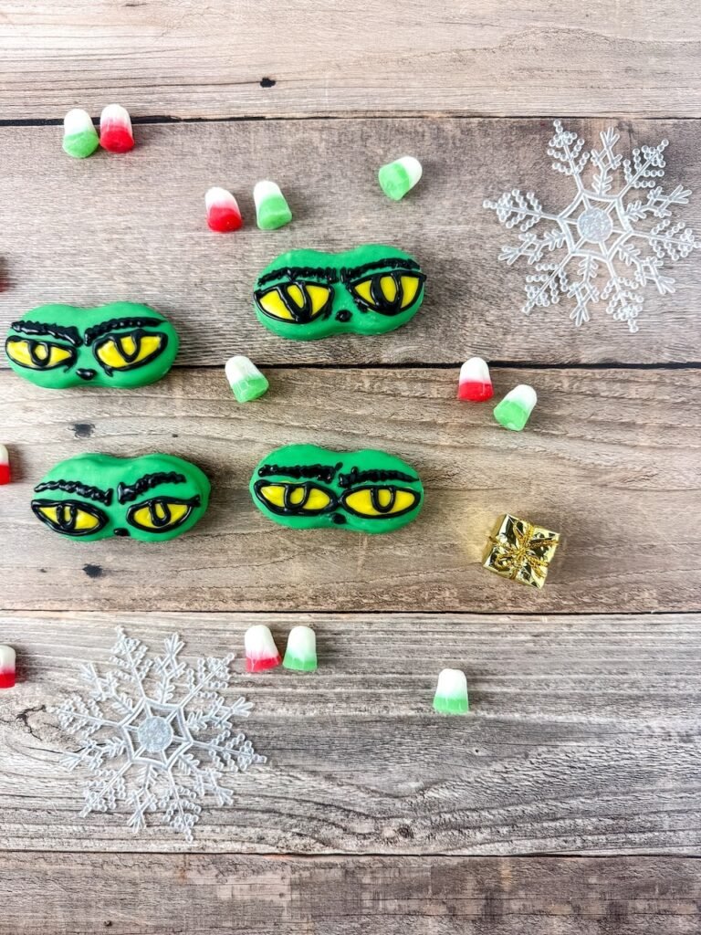 Overhead view of Grinch Nutter Butter cookies decorated with yellow eyes on a wooden table surrounded by red and green candies and snowflake decorations.