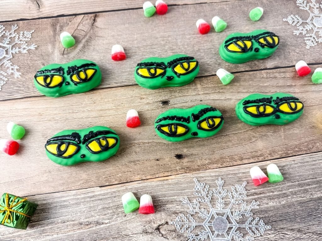 Grinch Nutter Butter cookies decorated with yellow eyes and black eyebrows on a wooden table with Christmas candy and snowflakes.