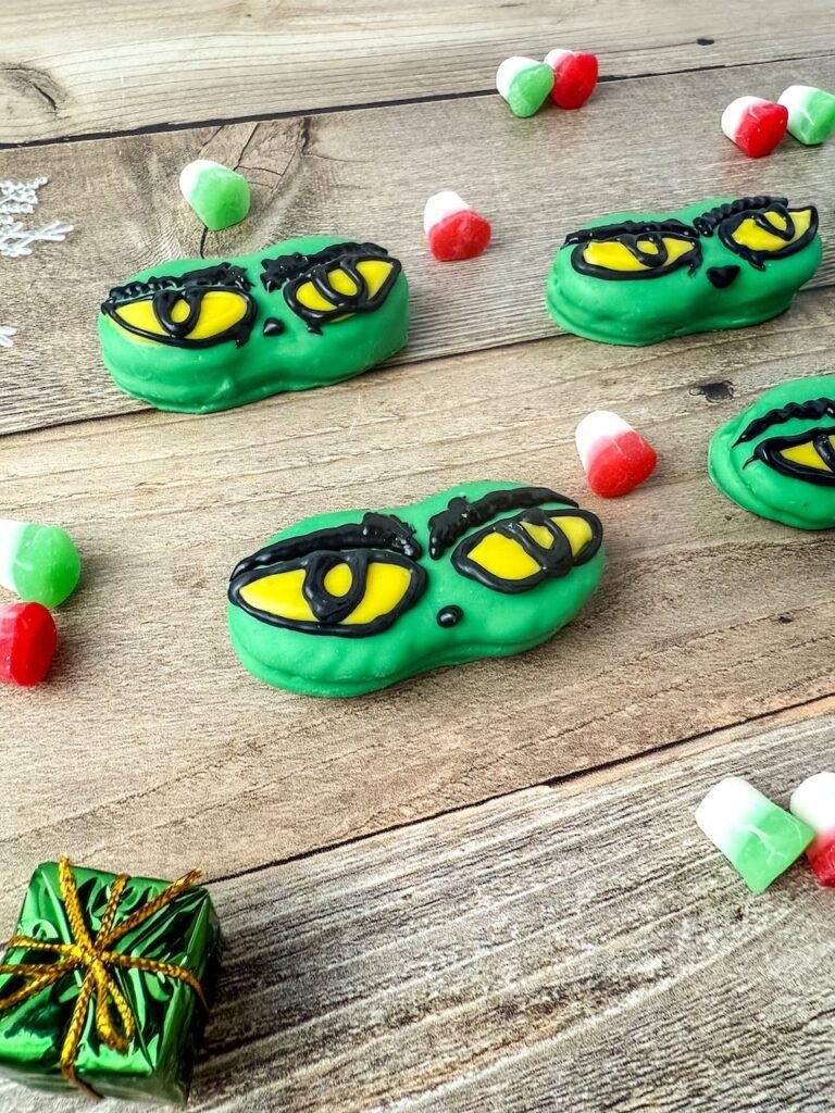 Festive Grinch Nutter Butter cookies decorated with yellow eyes and black eyebrows on a wooden table with Christmas candies.