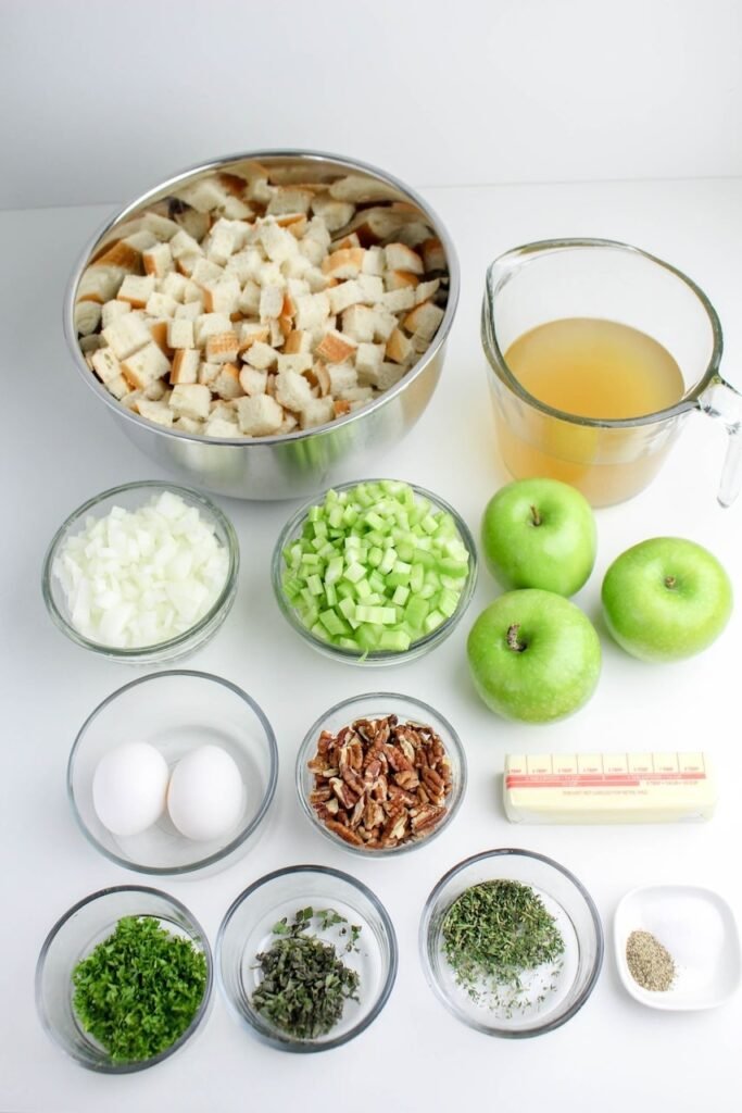 Ingredients for homemade herb and apple stuffing including bread cubes, green apples, celery, onion, eggs, herbs, pecans, butter, and broth arranged on a white surface.