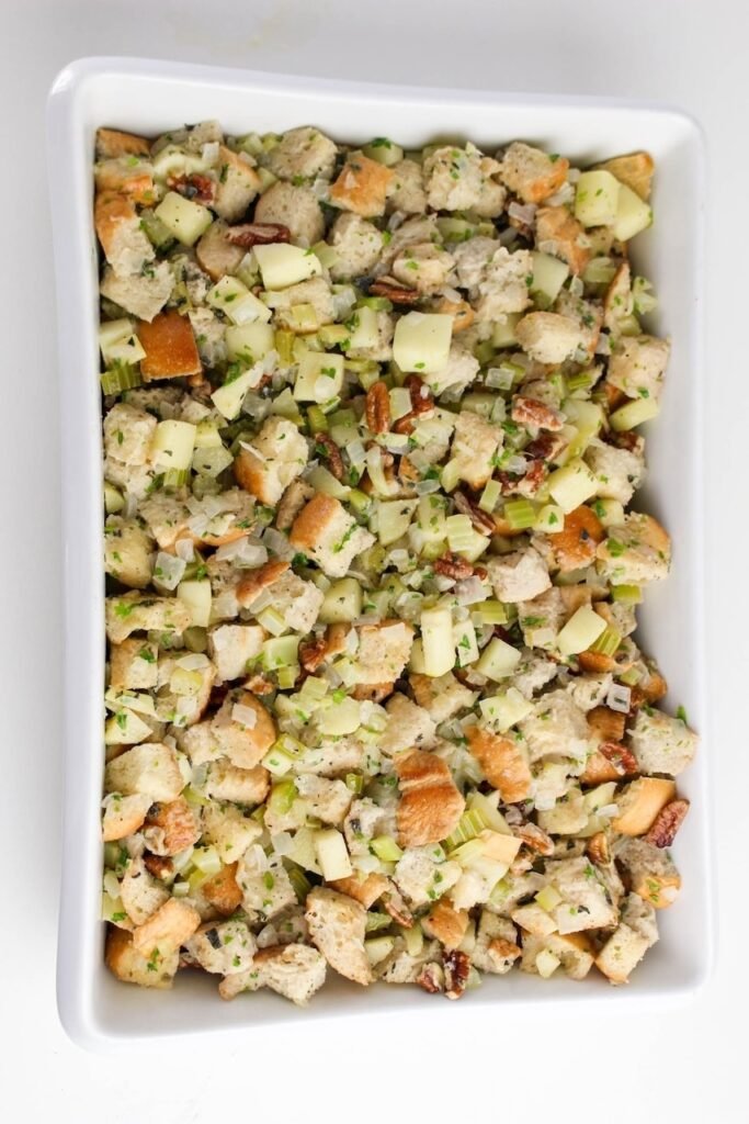 Unbaked herb and apple stuffing mixture with bread cubes, apples, celery, onions, and pecans spread evenly in a white baking dish.