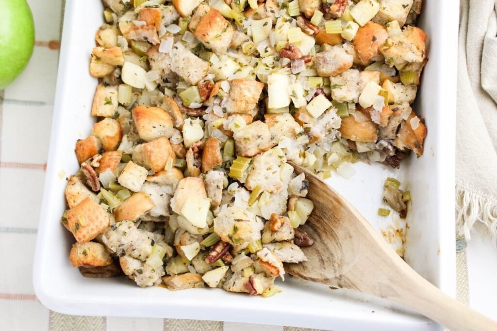 Partially served baked herb and apple stuffing in a white dish with a wooden spoon, showing golden bread cubes, apples, celery, onions, and pecans.