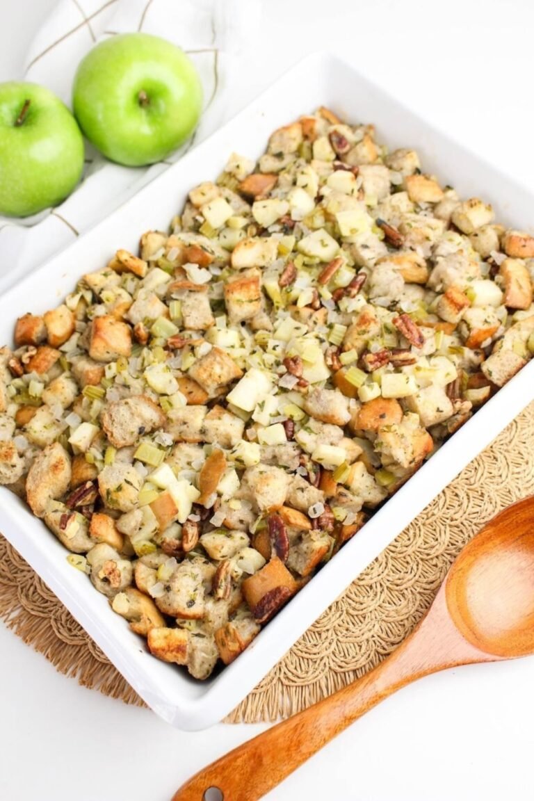 Golden baked herb and apple stuffing in a white dish, made with bread cubes, apples, celery, onions, and pecans, styled with green apples and a wooden spoon.