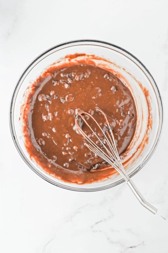 Chocolate brownie batter with chocolate chips being mixed in a glass bowl using a whisk during step two of making Grinch Brownies.