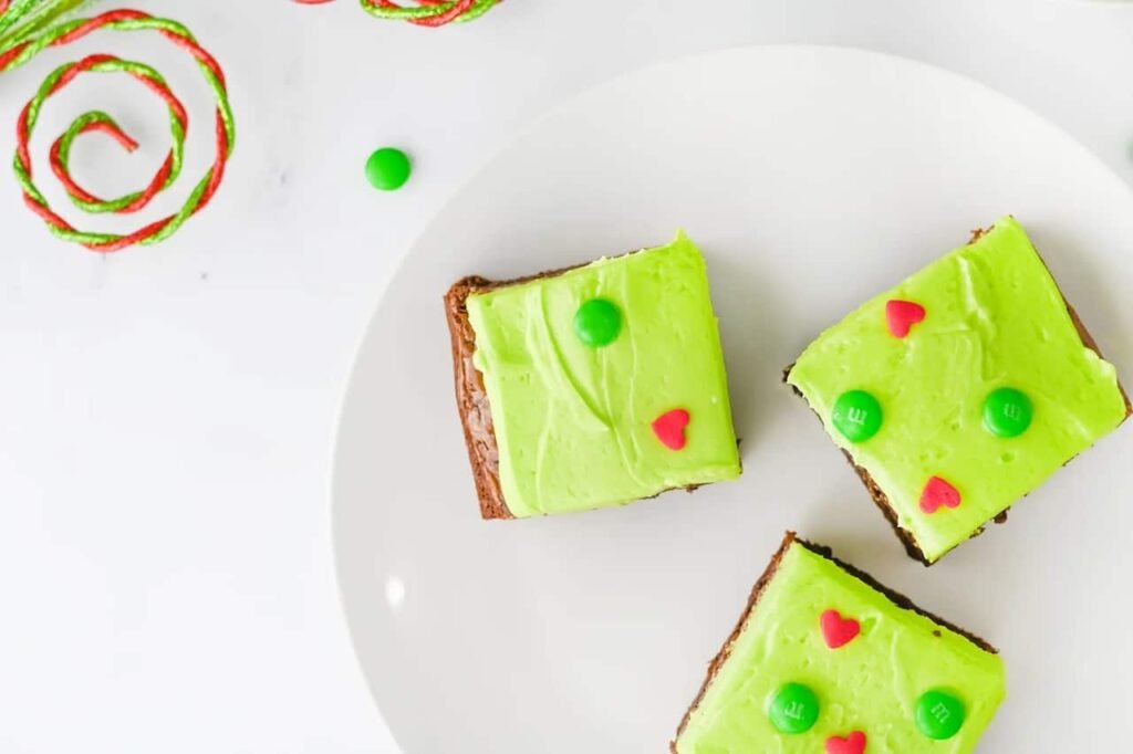 Overhead view of three Grinch Brownies topped with green frosting, red heart sprinkles, and green M&Ms on a white plate, surrounded by festive red and green ribbon.