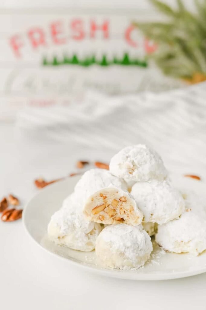 Stack of holiday pecan snowball cookies coated in powdered sugar on a white plate, surrounded by pecans with festive Christmas decor in the background.