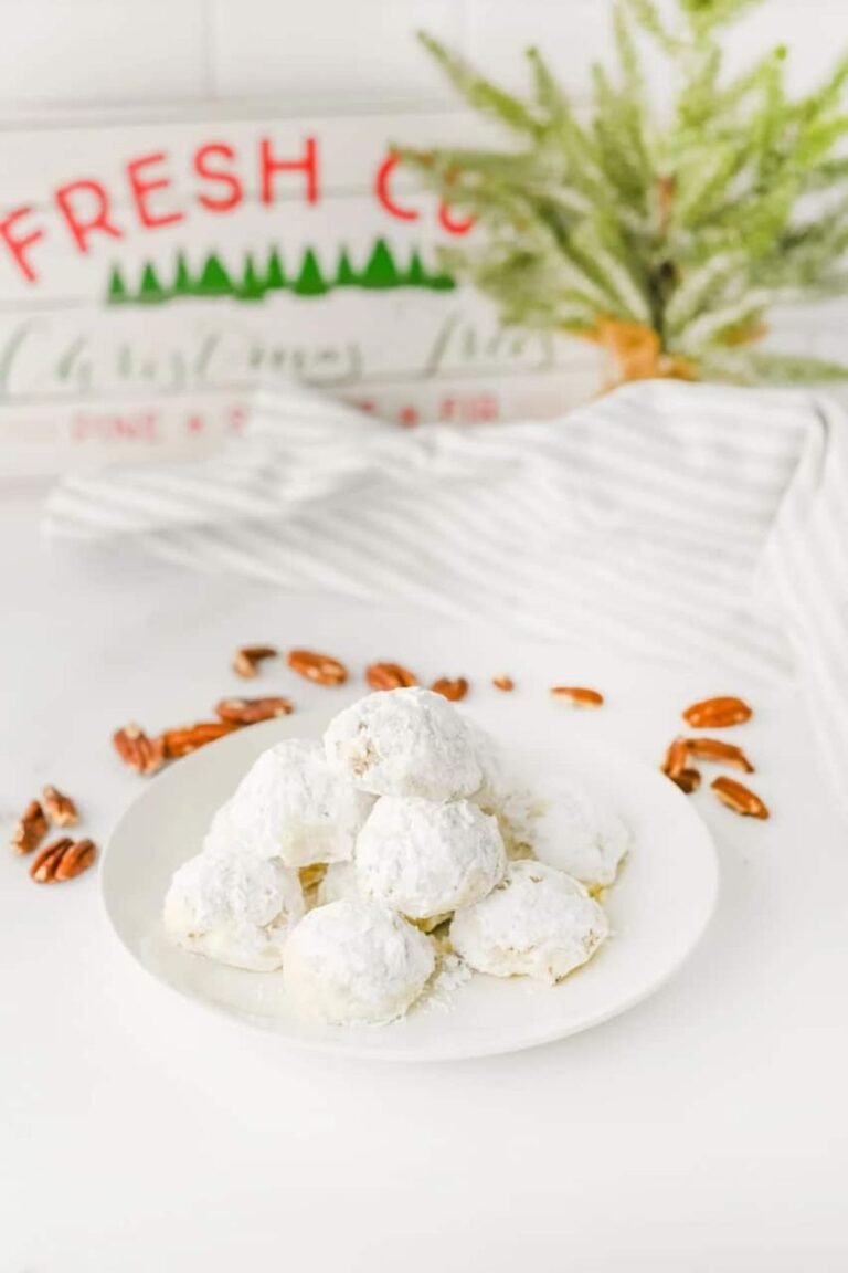 Plate of holiday pecan snowball cookies coated in powdered sugar, surrounded by pecans and festive Christmas decor with a “Fresh Cut Christmas Trees” sign in the background.