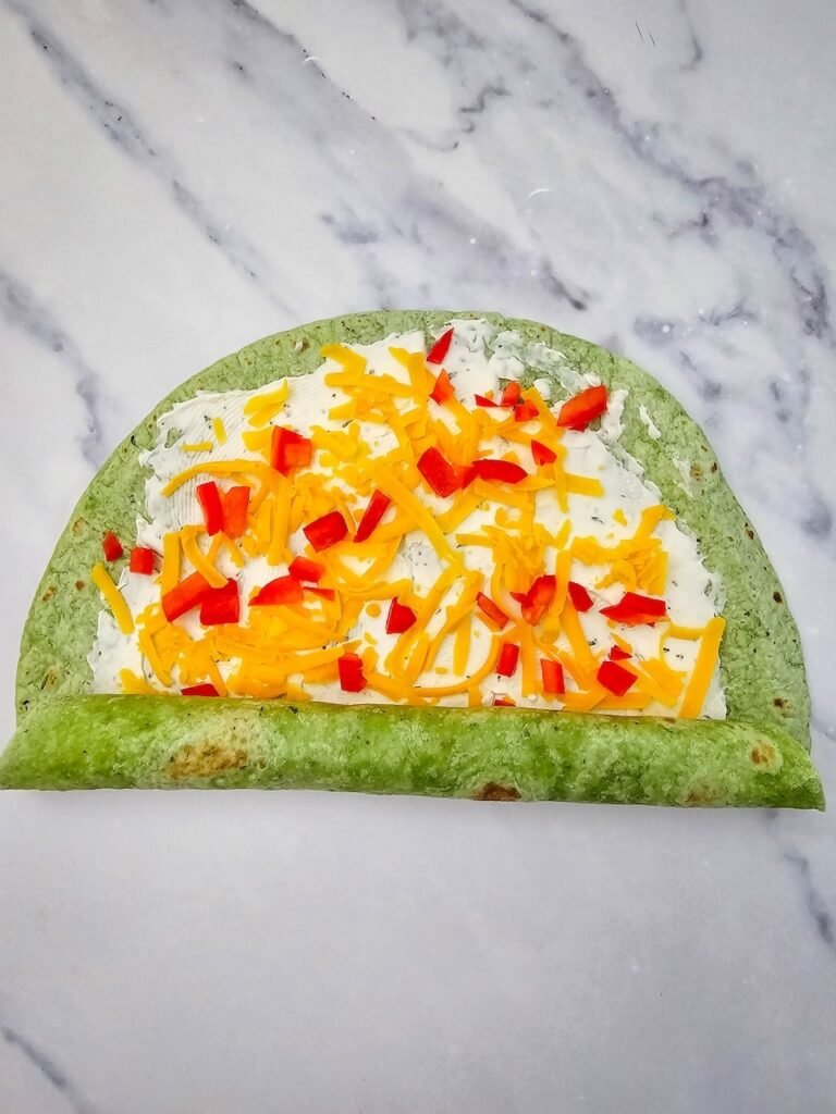 Spinach tortilla being rolled tightly on a marble surface after being spread with cream cheese and ranch seasoning, then topped with shredded cheddar cheese and diced red peppers. This step shows how to roll before wrapping in plastic and chilling for the holiday appetizer.