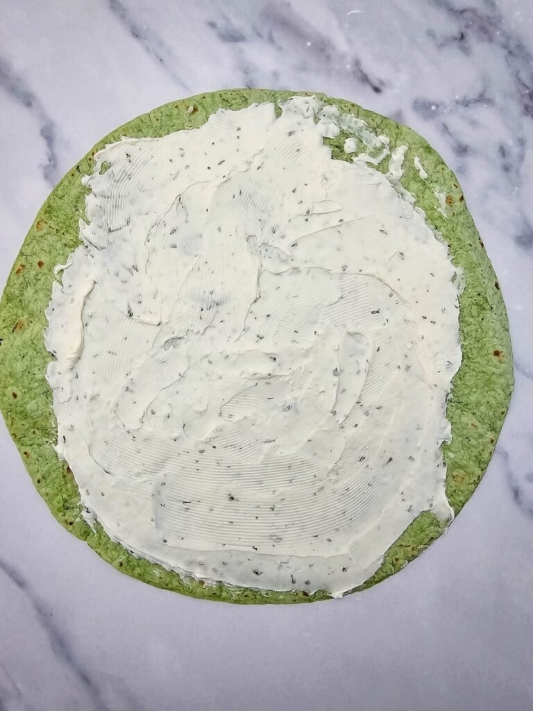 Spinach tortilla laid flat on a marble surface with a layer of softened cream cheese mixed with ranch seasoning spread evenly across the surface. This is the first step in assembling before adding shredded cheese and diced red pepper for the festive holiday appetizer.