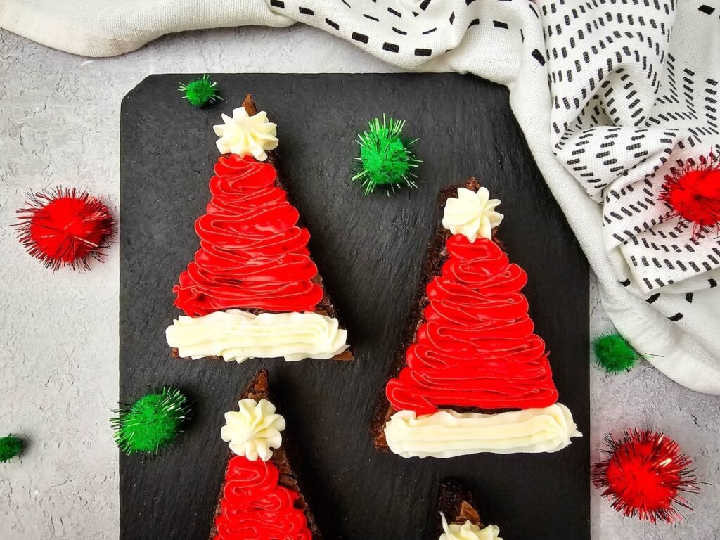 Close-up of decorated Santa Hat Brownies with red and white frosting on a black slate board surrounded by festive pom-poms.