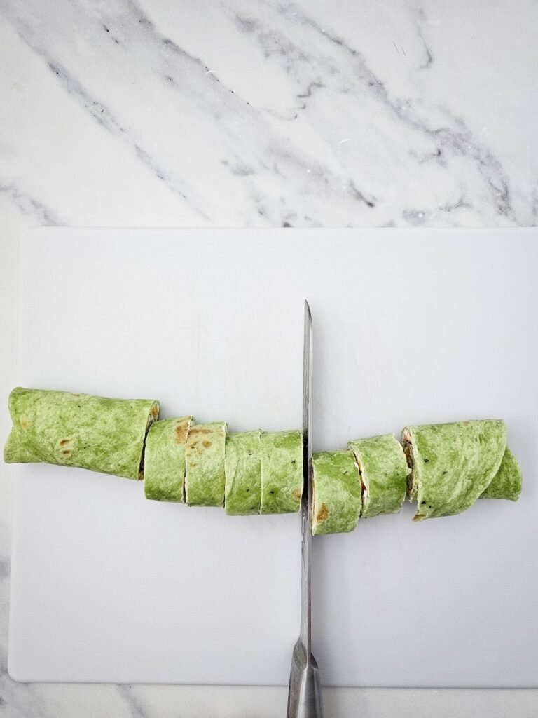 Spinach tortilla roll filled with cream cheese, cheddar cheese, and diced red peppers being sliced with a serrated knife on a white cutting board. This step shows how to cuti even pieces before arranging them into a festive holiday appetizer tree.