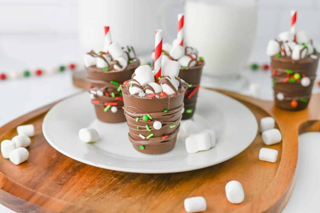Homemade Christmas hot chocolate sticks decorated with mini marshmallows and festive sprinkles, served on a white plate with milk in the background.