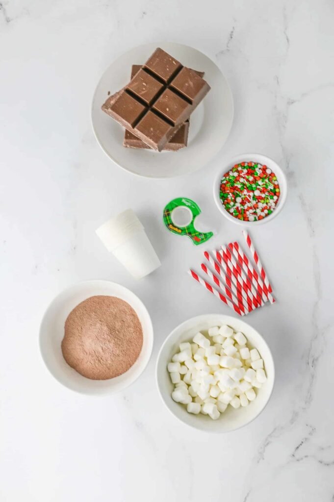 Flat lay of ingredients for homemade hot chocolate sticks, including chocolate, cocoa mix, mini marshmallows, straws, and festive sprinkles on a marble background.