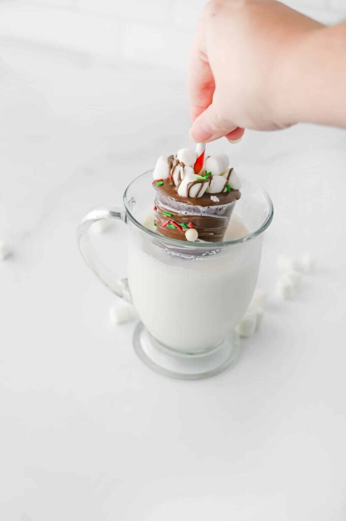 Hand dipping a homemade hot chocolate stick topped with marshmallows into a mug of warm milk to make creamy hot cocoa.