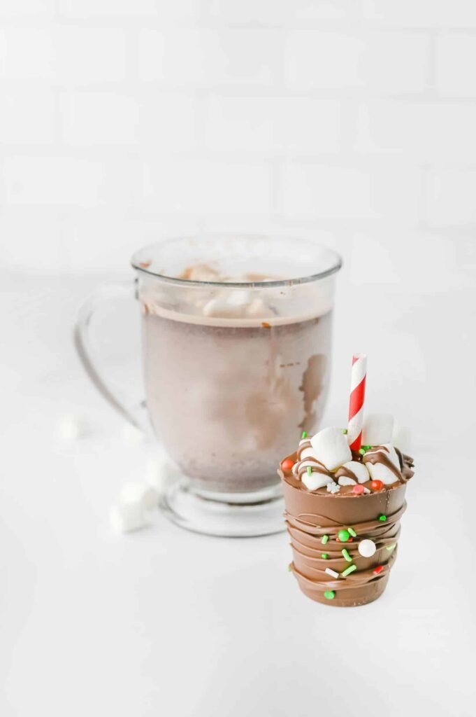 Homemade hot chocolate sticks with red striped straws beside a glass mug of creamy hot cocoa topped with marshmallows.