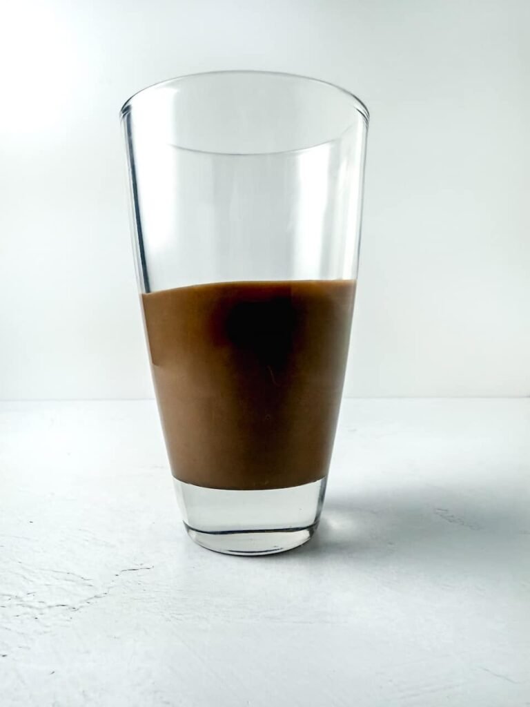 Step three showing a tall glass partially filled with smooth melted chocolate, ready for dipping pretzel rods. The glass is placed on a clean white surface to demonstrate the dipping step for coating the pretzels evenly.